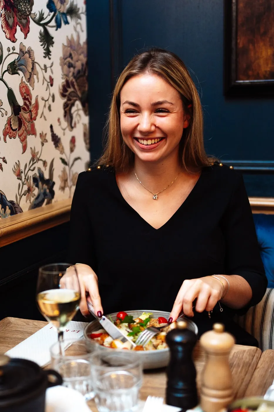 Smiling woman in black dress sitting at a restaurant table, cutting a salad with fork and knife.