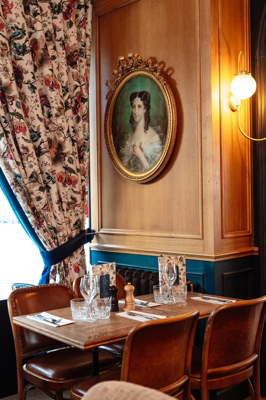 Cozy restaurant dining corner with a wooden table set for four, leather chairs, floral curtains, and a vintage portrait on a wood-paneled wall.