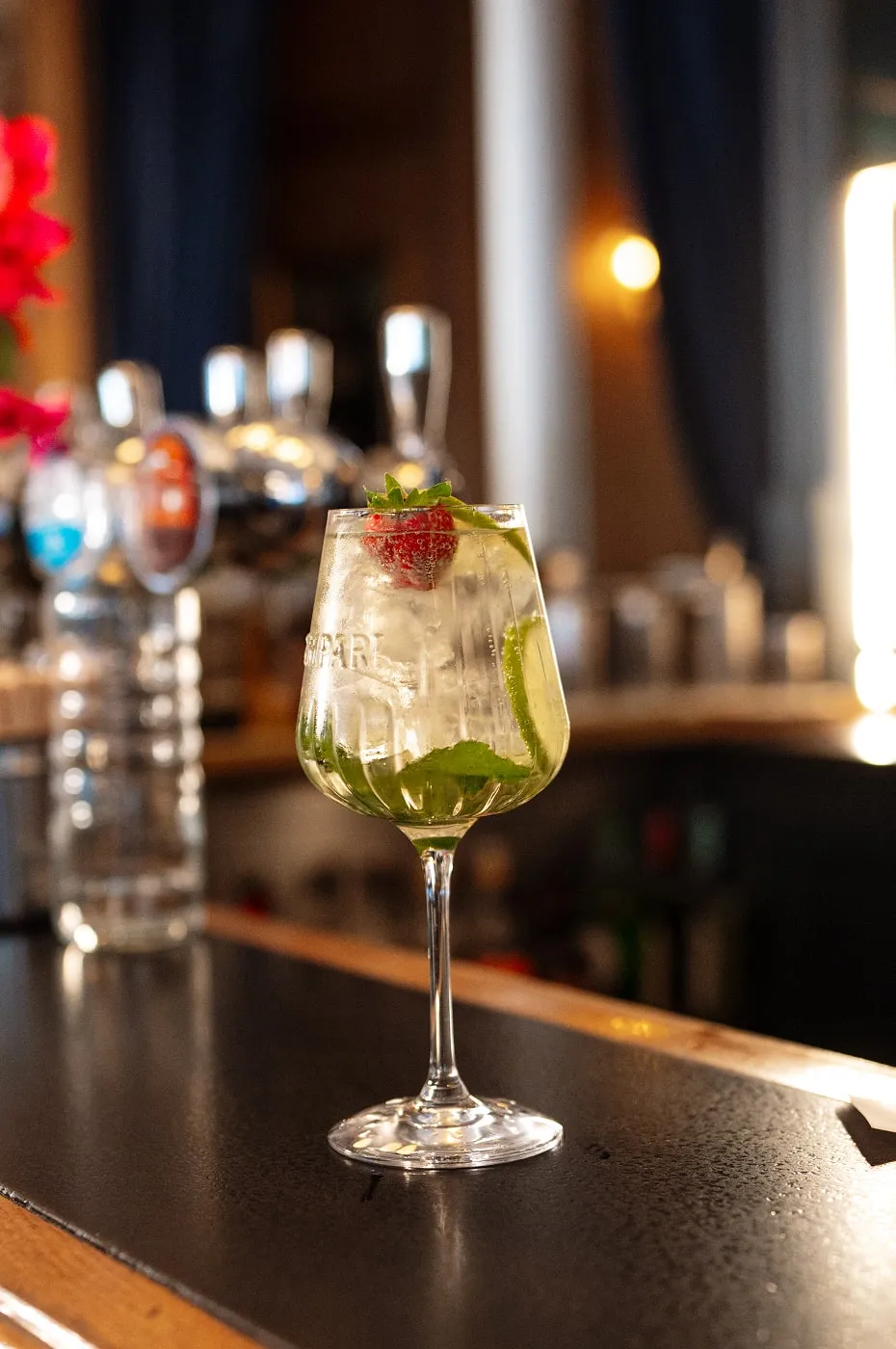 Cocktail glass with ice, mint leaves, lime slice, and a strawberry garnish on a bar counter.
