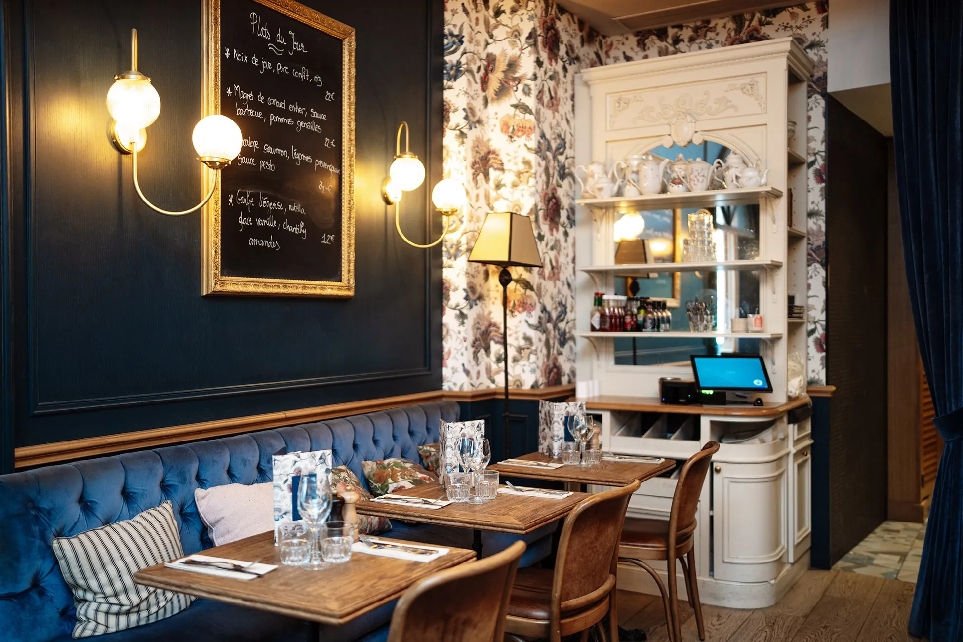 Cozy restaurant corner with wooden tables set for dining, blue tufted bench seating, floral wallpaper, and a framed chalkboard menu.