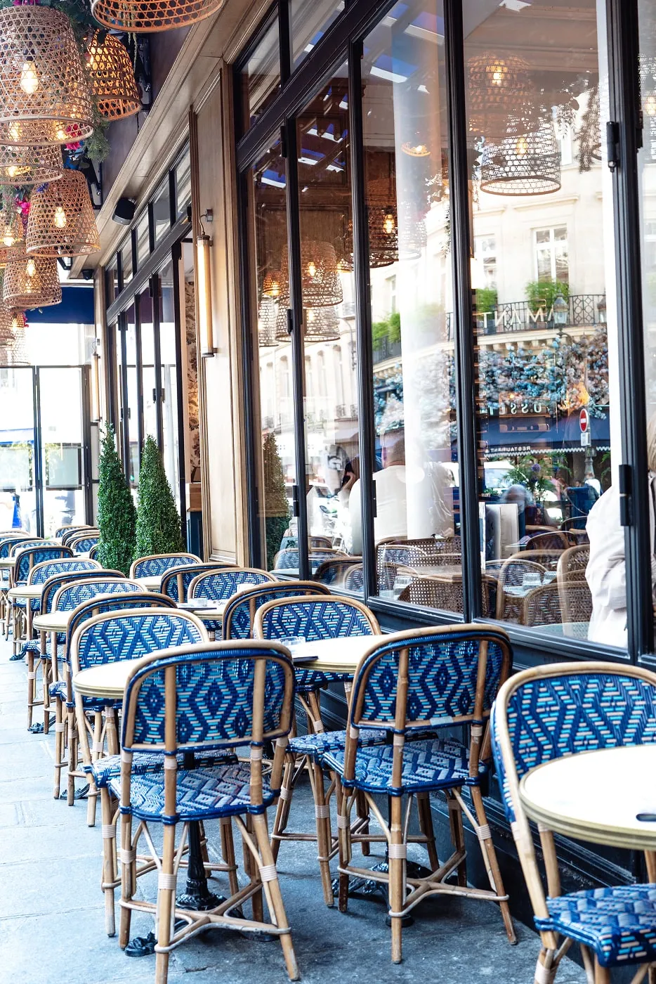 Outdoor café seating with blue patterned chairs and round tables along a sidewalk next to large glass windows reflecting street scene.