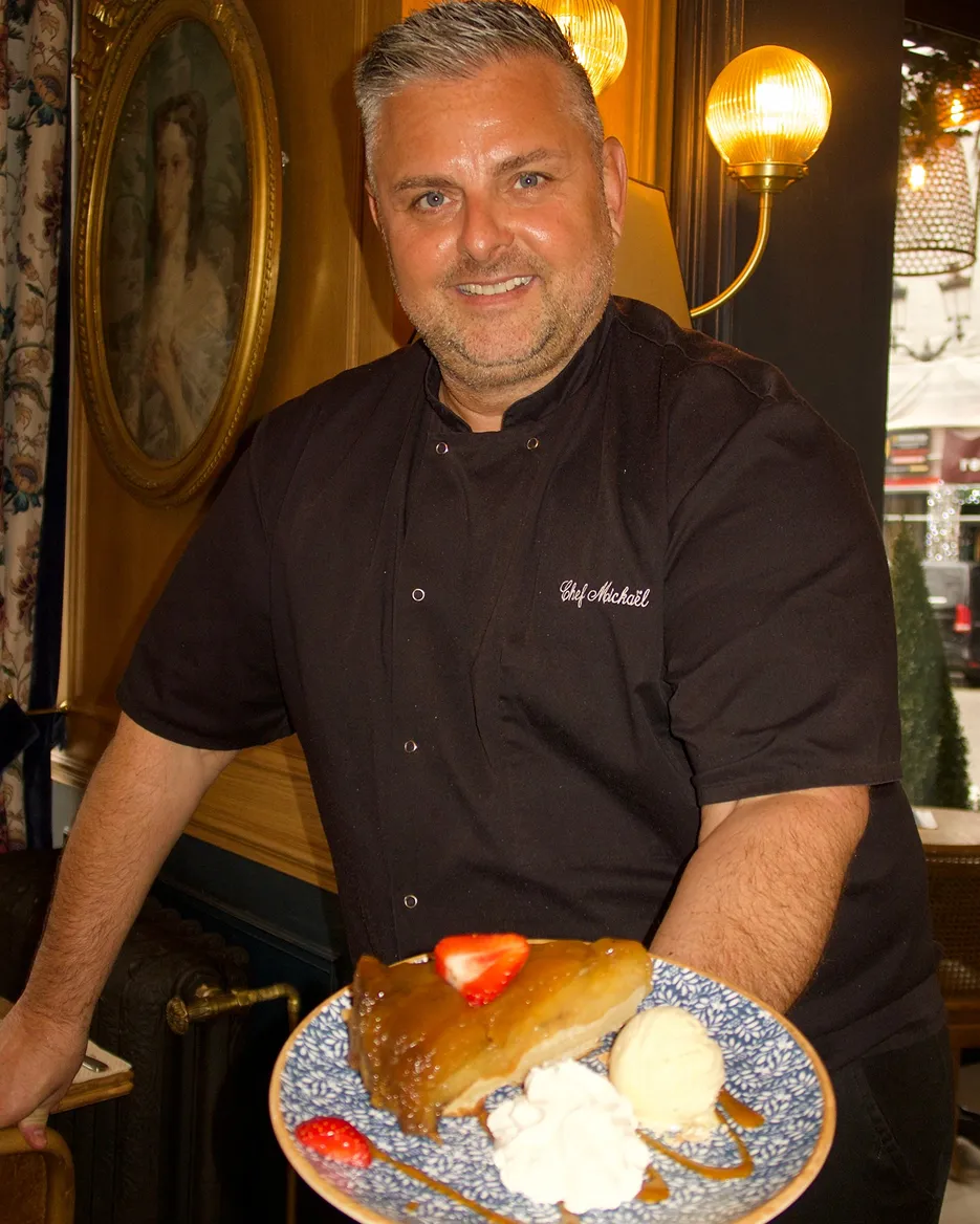 Chef in black uniform holding a plate with dessert topped with strawberries, whipped cream, and ice cream inside a warmly lit restaurant.