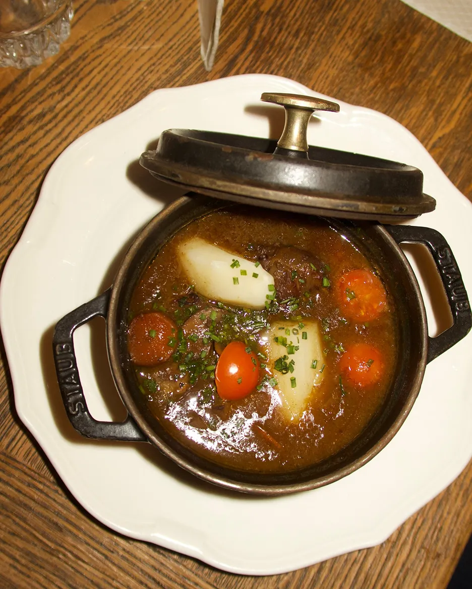 Cast iron pot with stew containing carrots, potatoes, meat, and garnished with chopped herbs on a white plate over a wooden table.