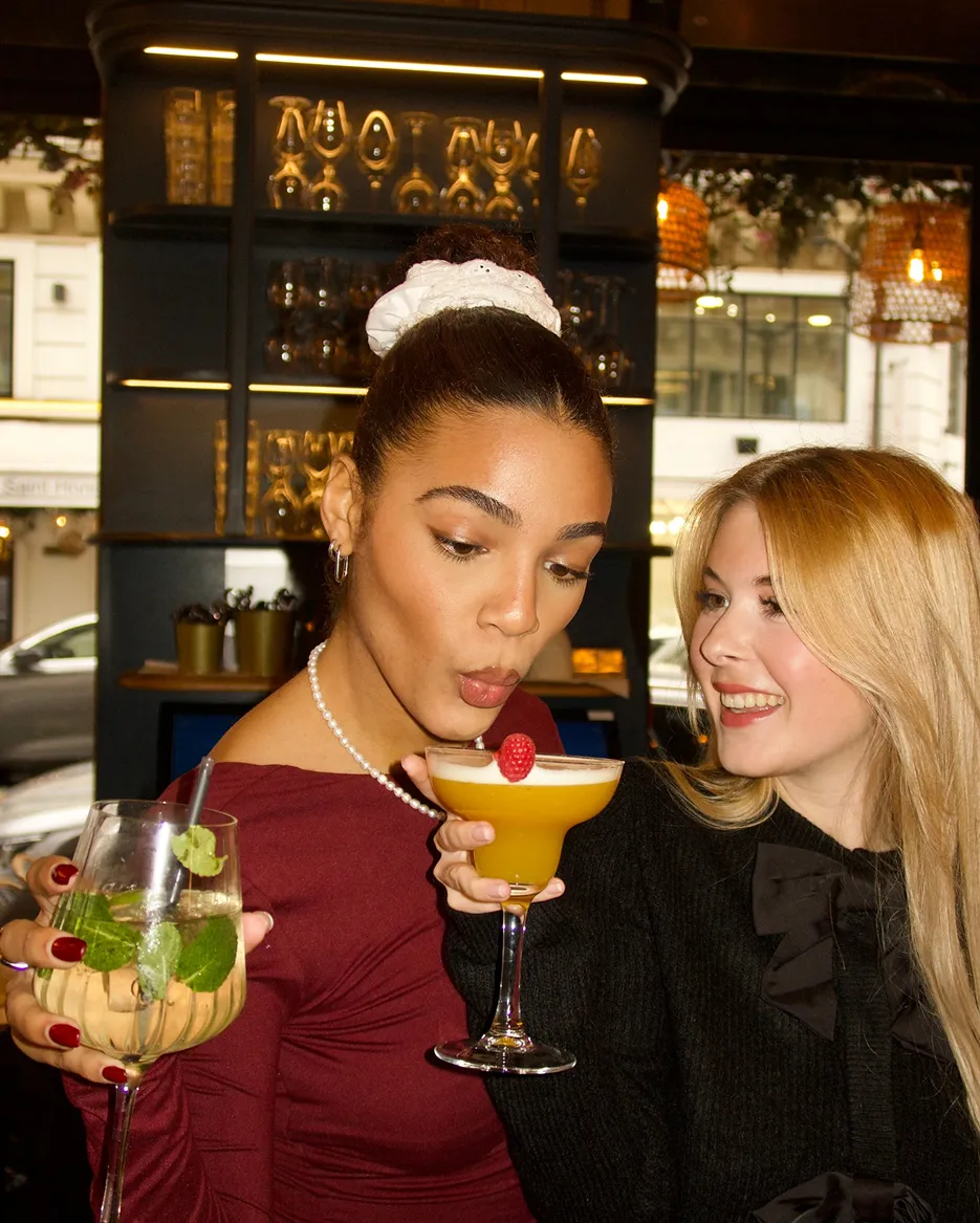 Two women smiling and enjoying cocktails at a bar, one holding a yellow drink with a raspberry garnish and the other holding a glass with mint leaves.