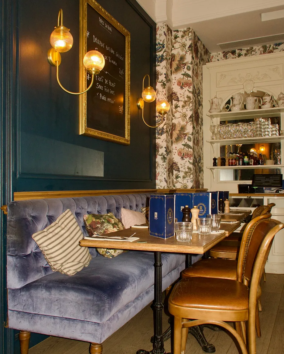 Cozy dining area with blue velvet bench, wooden chairs, hanging sconces, and shelves with glassware and ceramics.
