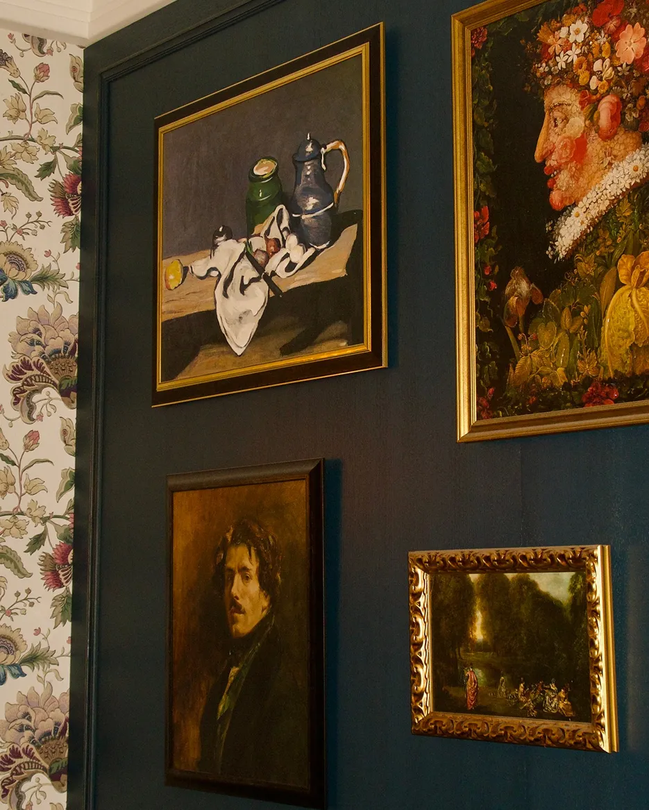 Four framed paintings on a dark blue wall next to floral wallpaper, including a still life, a portrait, a floral face artwork, and a small landscape scene.