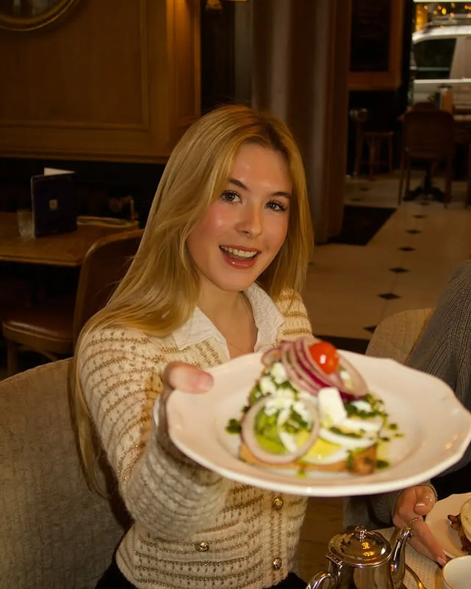 Smiling woman with long blonde hair holding a plate of salad with onions, cherry tomato, and feta cheese in a cozy restaurant.