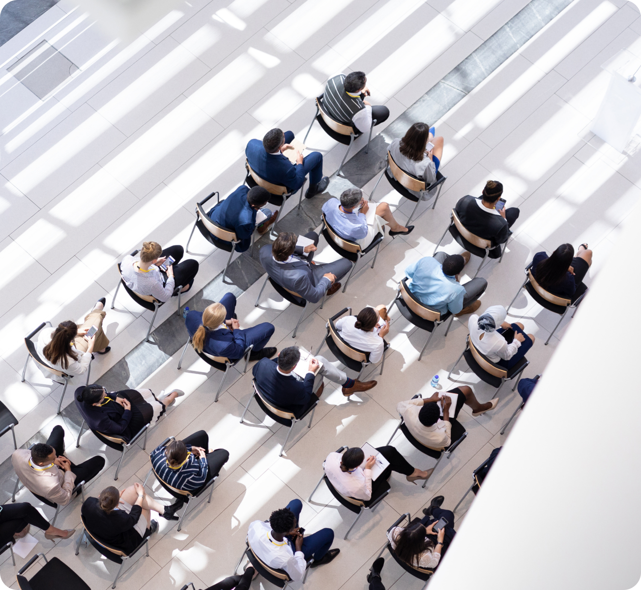 Overhead view of a group of people sitting in spaced chairs, some using phones or taking notes, in a bright, modern indoor space.