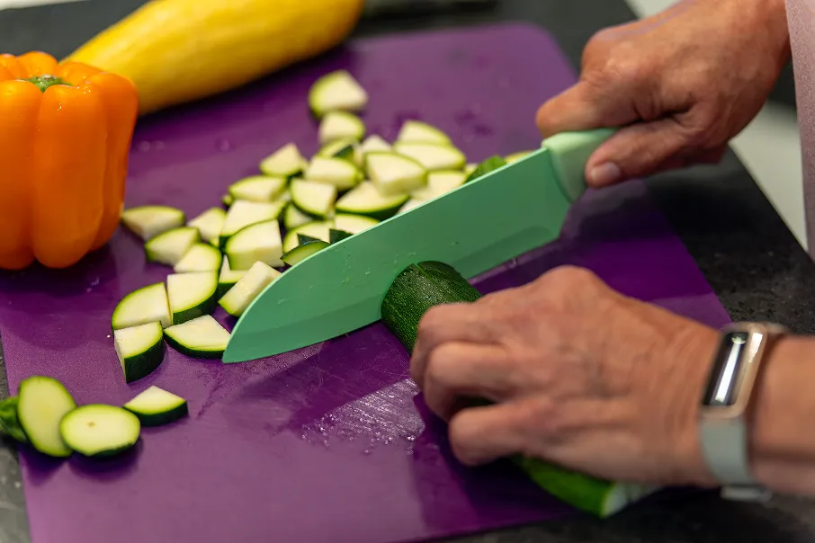 Hands with a wristwatch chopping a green zucchini on a purple cutting board with sliced zucchini and an orange bell pepper nearby.