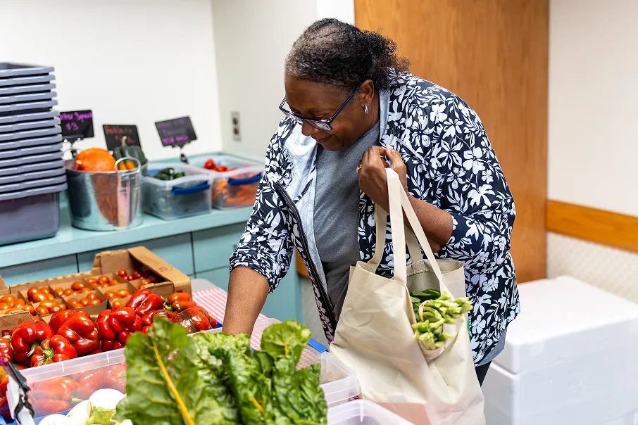 Woman smiling and holding a reusable shopping bag filled with fresh vegetables while selecting produce at an indoor market.