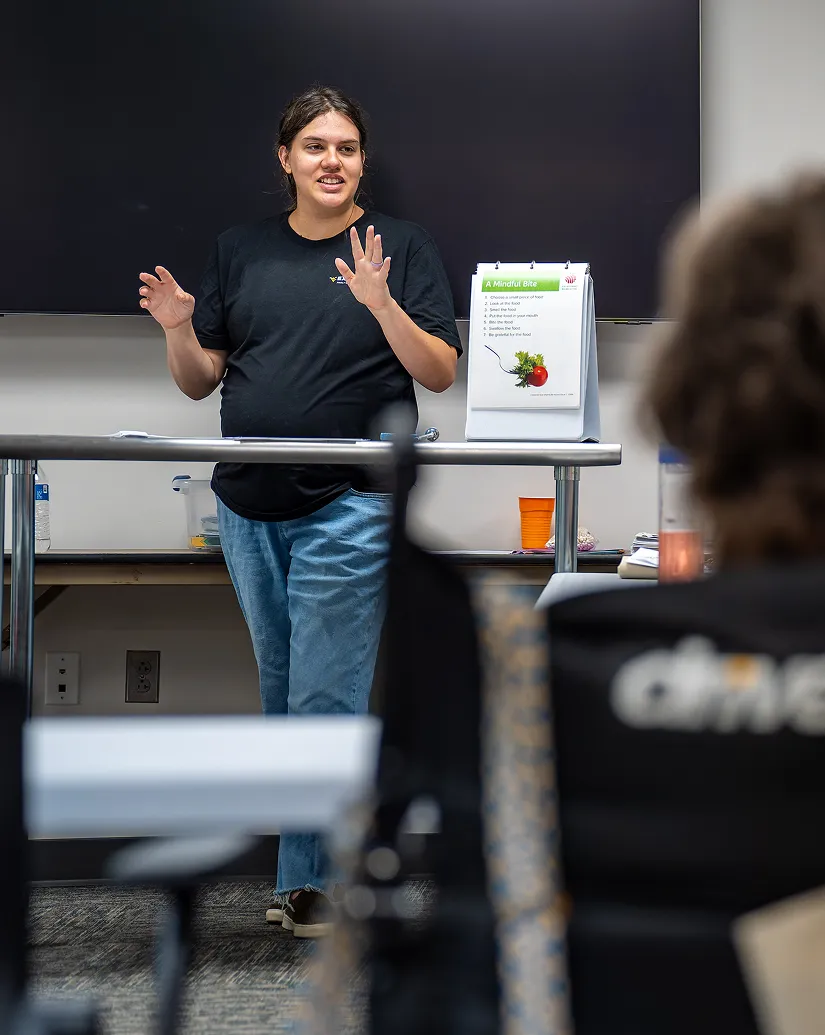 Woman in a black t-shirt and jeans speaking in front of a classroom with a large screen behind her and a flip chart titled 'A Mindful Bite'.