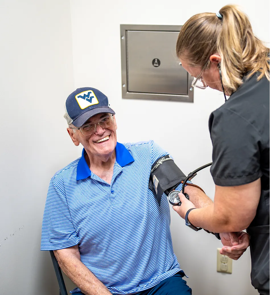 Smiling elderly man wearing a blue striped shirt and West Virginia cap having his blood pressure measured by a healthcare worker.