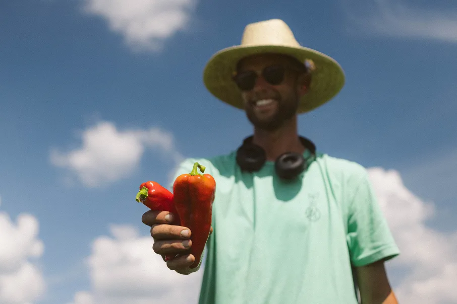 Man in a straw hat and sunglasses holding two red bell peppers against a blue sky with clouds.