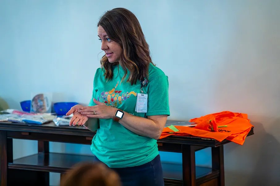 Woman in a green shirt giving a presentation or talking indoors near a table with folded clothes and orange safety vests.