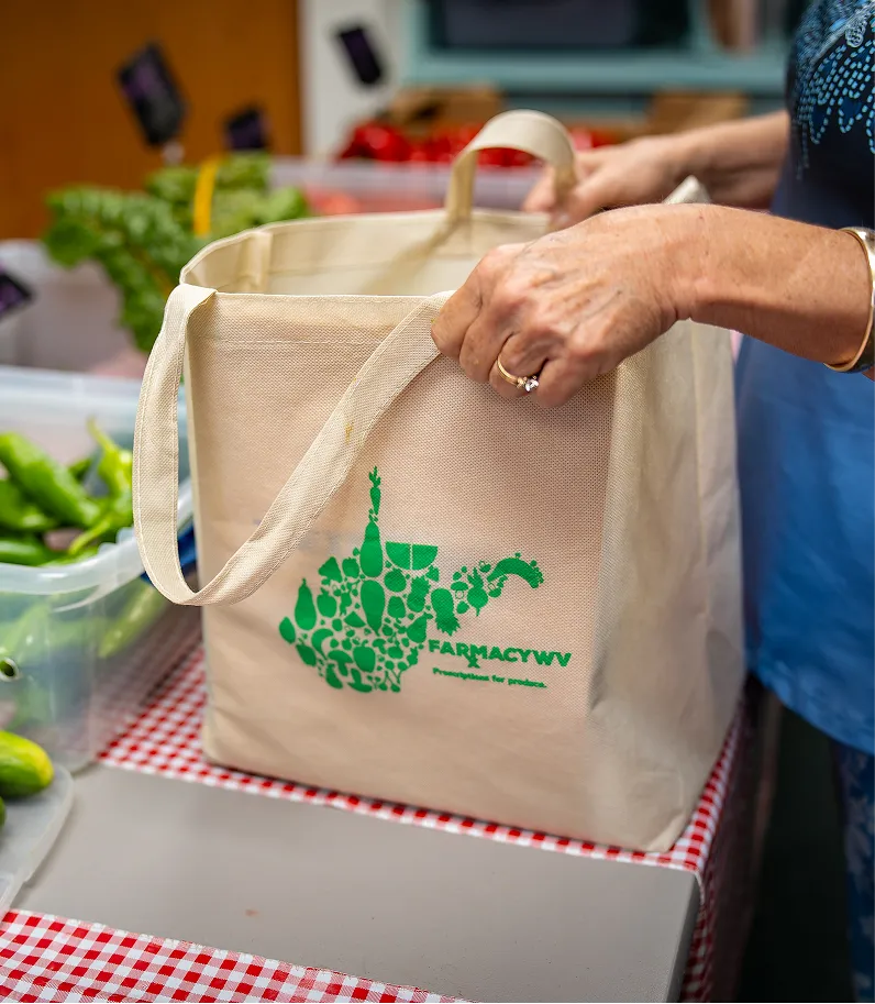 Person holding a beige FARMacyWV reusable shopping bag on a red and white checkered table, with fresh green vegetables in plastic containers nearby.