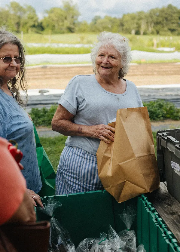 Smiling elderly woman holding a brown paper bag at an outdoor market while another woman stands nearby.