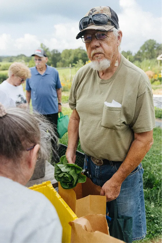Elderly man wearing a cap and sunglasses holds fresh greens at an outdoor market with other people in the background.