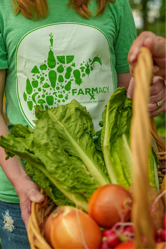 Person wearing a green Farmacy t-shirt holding a basket filled with fresh lettuce, onions, and radishes.
