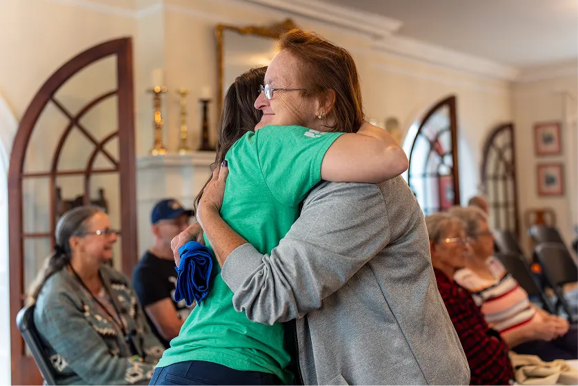 Two women warmly hugging inside a room, while other elderly people sit and chat in the background.