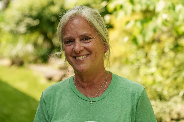 Smiling woman with blonde hair wearing a green shirt and necklace standing outdoors with blurred green foliage background.