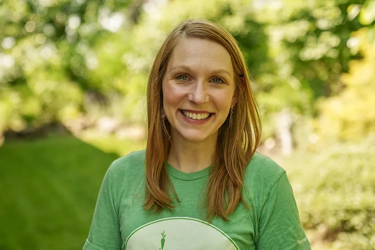 Smiling woman with long red hair wearing a green T-shirt standing outdoors with blurred green foliage in the background.