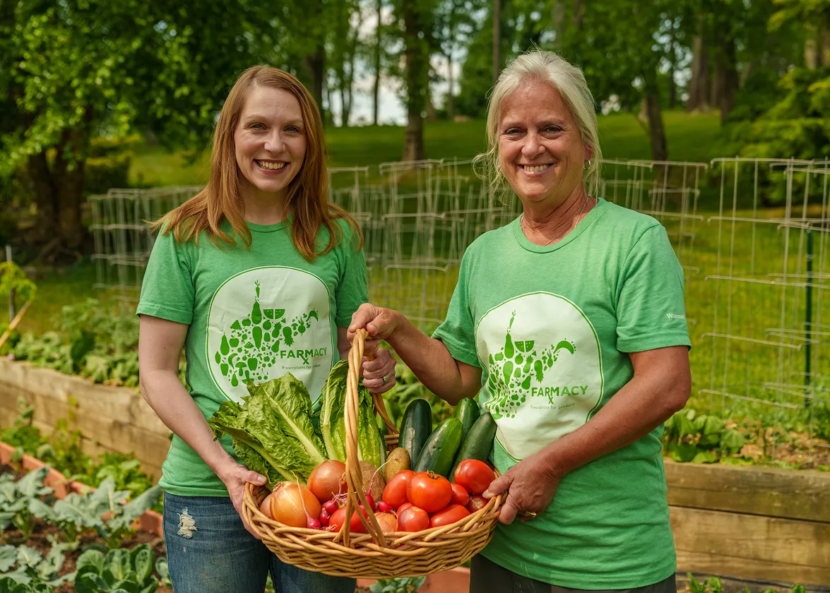 Two smiling women in green Farmacy t-shirts holding a basket of fresh vegetables in a garden.