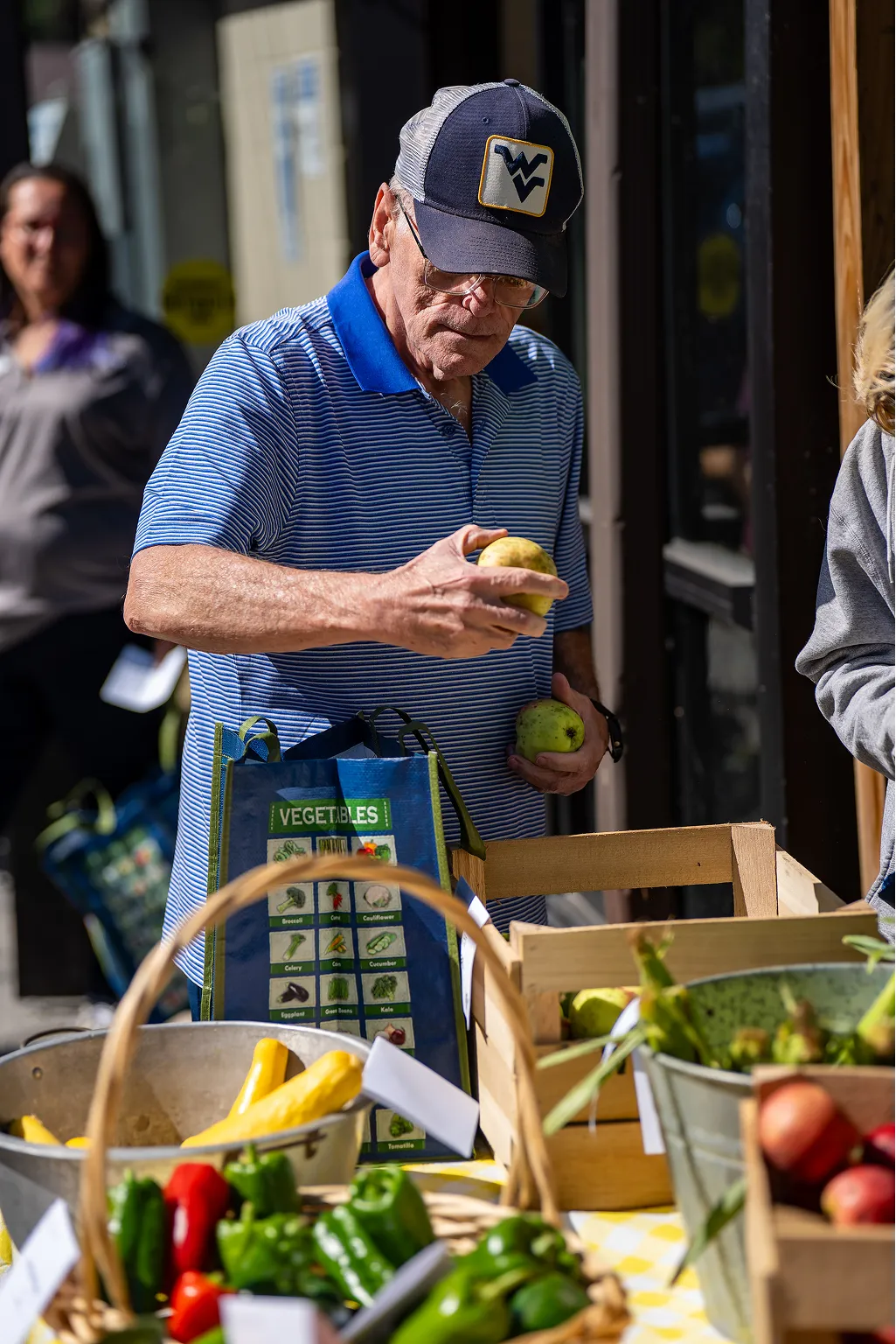 An elderly man in a striped blue polo shirt and a navy West Virginia cap selects apples at an outdoor market stall with baskets of vegetables and fruits.