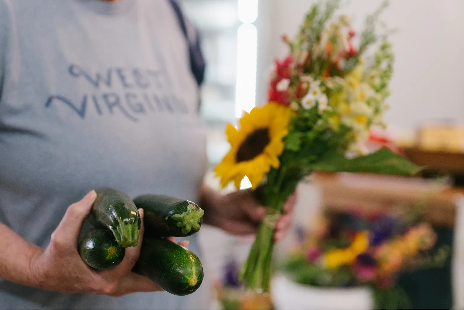 Person wearing a gray Sweet Virginia shirt holding four zucchinis in one hand and a bouquet of colorful flowers including a sunflower in the other.