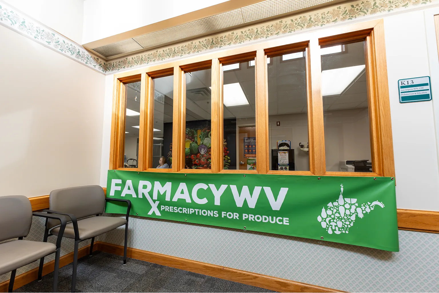 Waiting area with two chairs and a green banner reading 'FARMACYWV PRESCRIPTIONS FOR PRODUCE' featuring a West Virginia state silhouette made from fruit and vegetable icons.