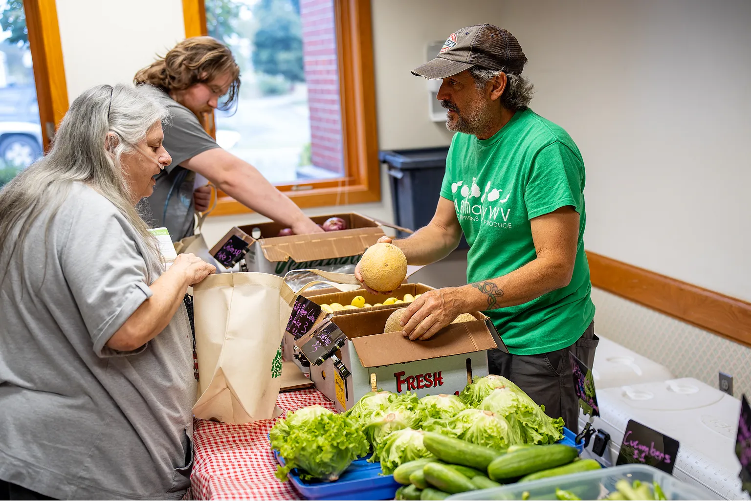 Man in green shirt with a cap handing a cantaloupe to a woman bagging groceries at a market table with fresh lettuce and cucumbers.