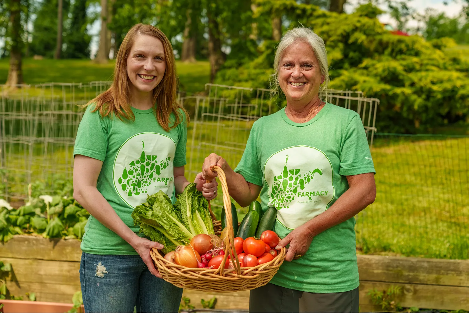 Two smiling women in green 'Farmacy' t-shirts holding a basket filled with fresh vegetables including lettuce, tomatoes, onions, and cucumbers in a garden.