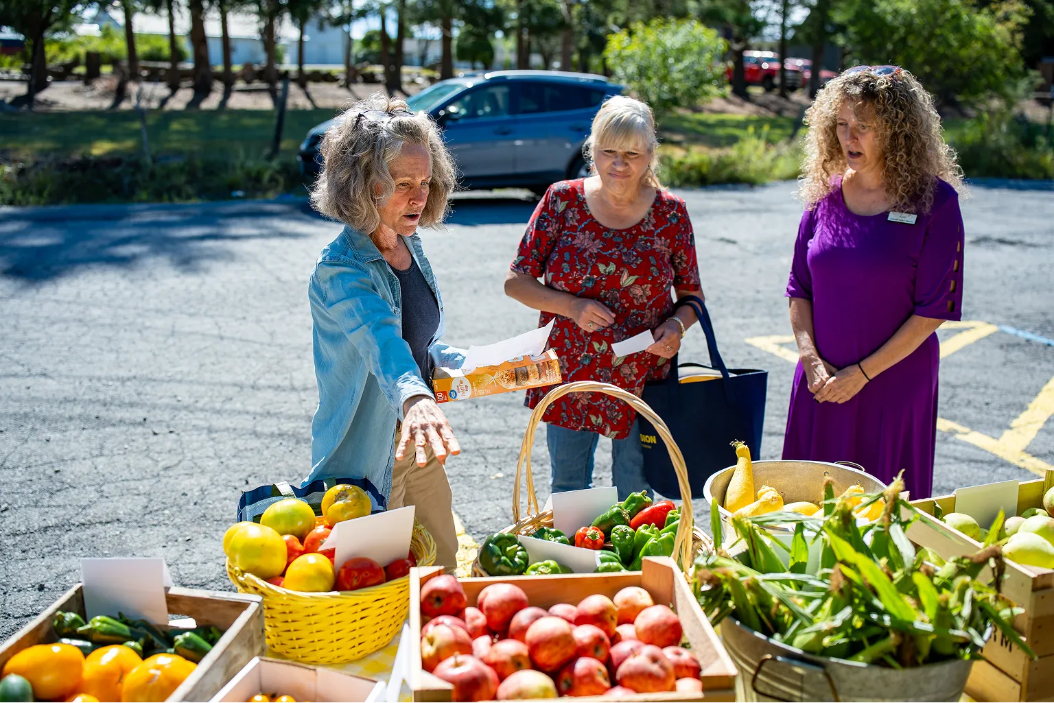 Three women shopping outdoors at a farmers market, selecting fresh produce like tomatoes, peppers, apples, corn, and squash from baskets and crates.