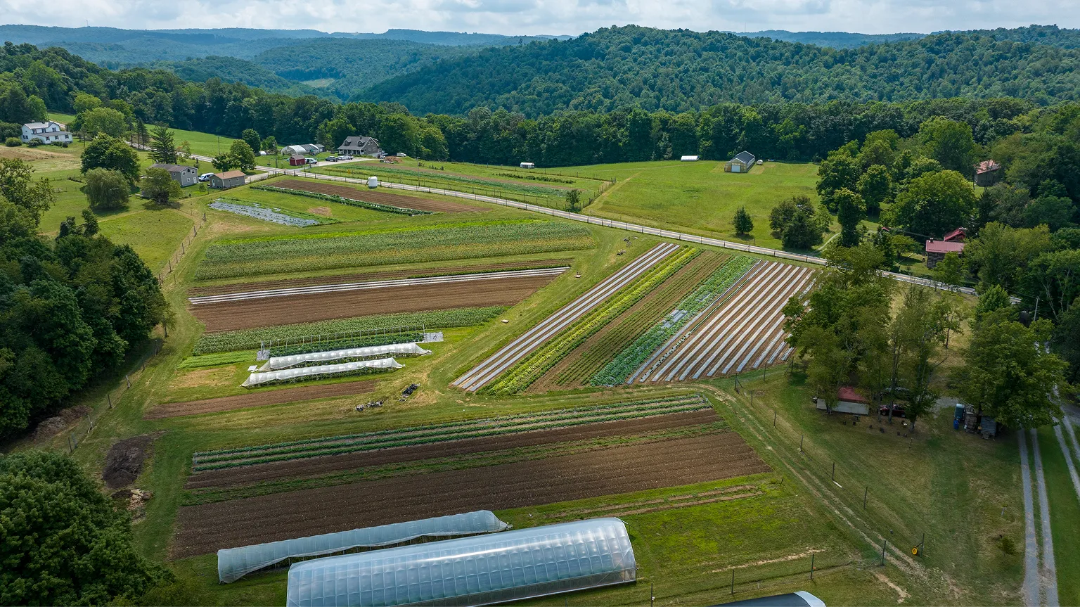 Aerial view of a farm with multiple crop fields, greenhouses, and houses surrounded by trees and hills under a partly cloudy sky.