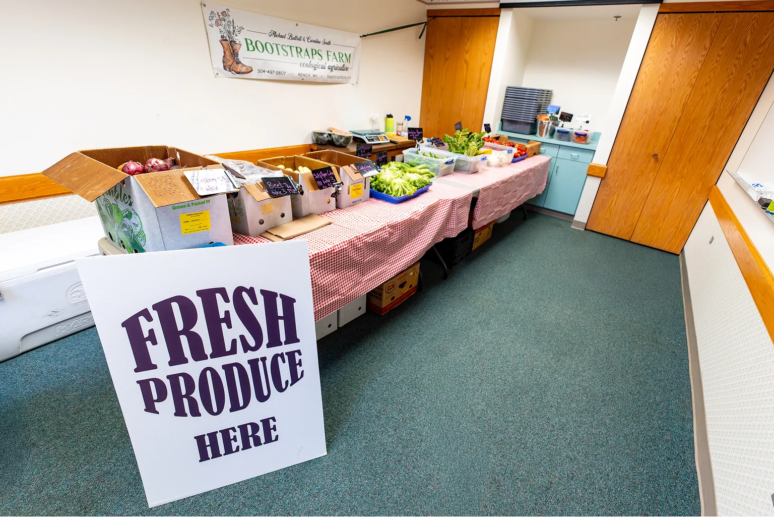 Indoor table with assorted fresh vegetables in boxes and containers, with a sign reading 'FRESH PRODUCE HERE' in front.