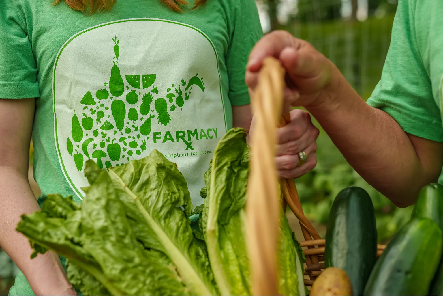 Person wearing a green Farmacy shirt holding a basket filled with fresh lettuce, cucumbers, and potatoes.