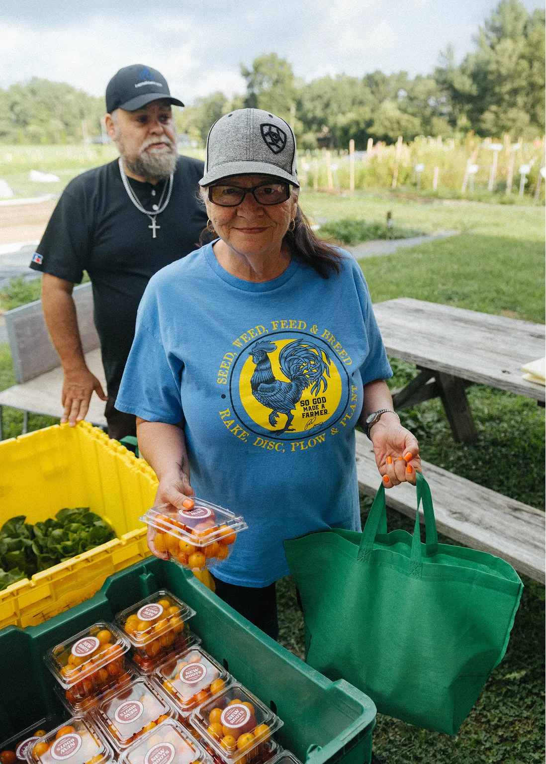 Smiling woman in a blue farmer-themed t-shirt and gray cap holding a container of cherry tomatoes and a green tote bag, with a man standing behind near crates of produce outdoors.