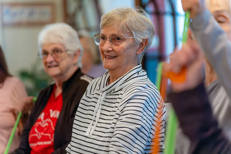 Smiling elderly woman with glasses and short grey hair wearing a white and black striped hoodie during a group activity.