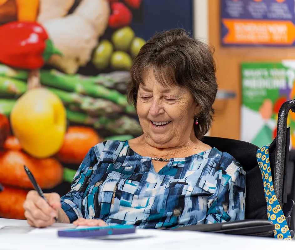 Smiling elderly woman in a patterned blue top writing with a pen at a table, with a colorful produce background.