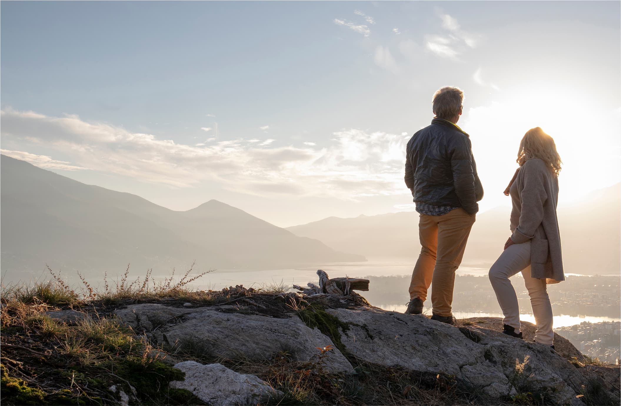 "Two people stand on rocky outcrop overlooking misty mountain landscape at golden hour, with layered mountain silhouettes fading into the distance under a cloudy sky