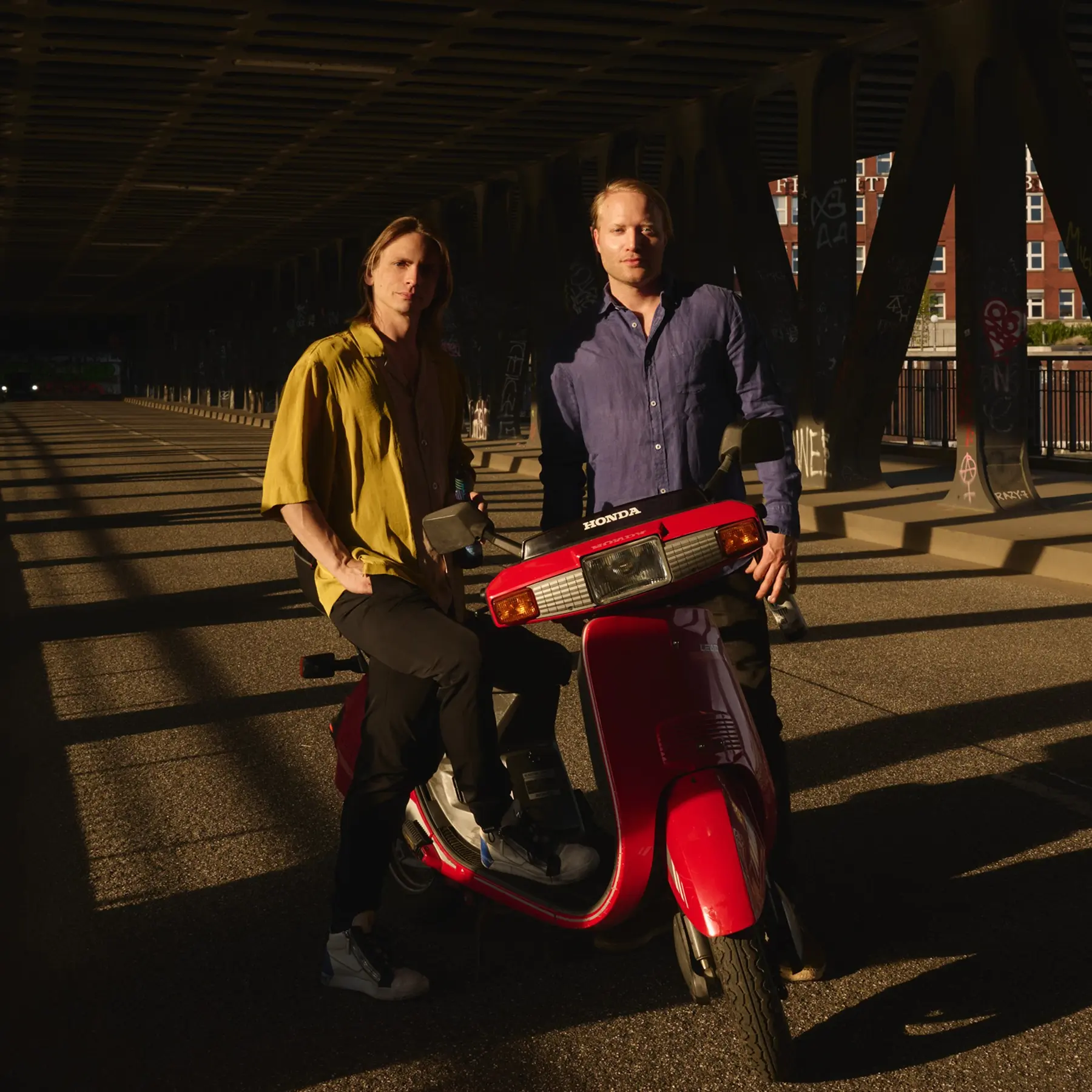 Two men posing with a red Honda scooter under an urban bridge at golden hour