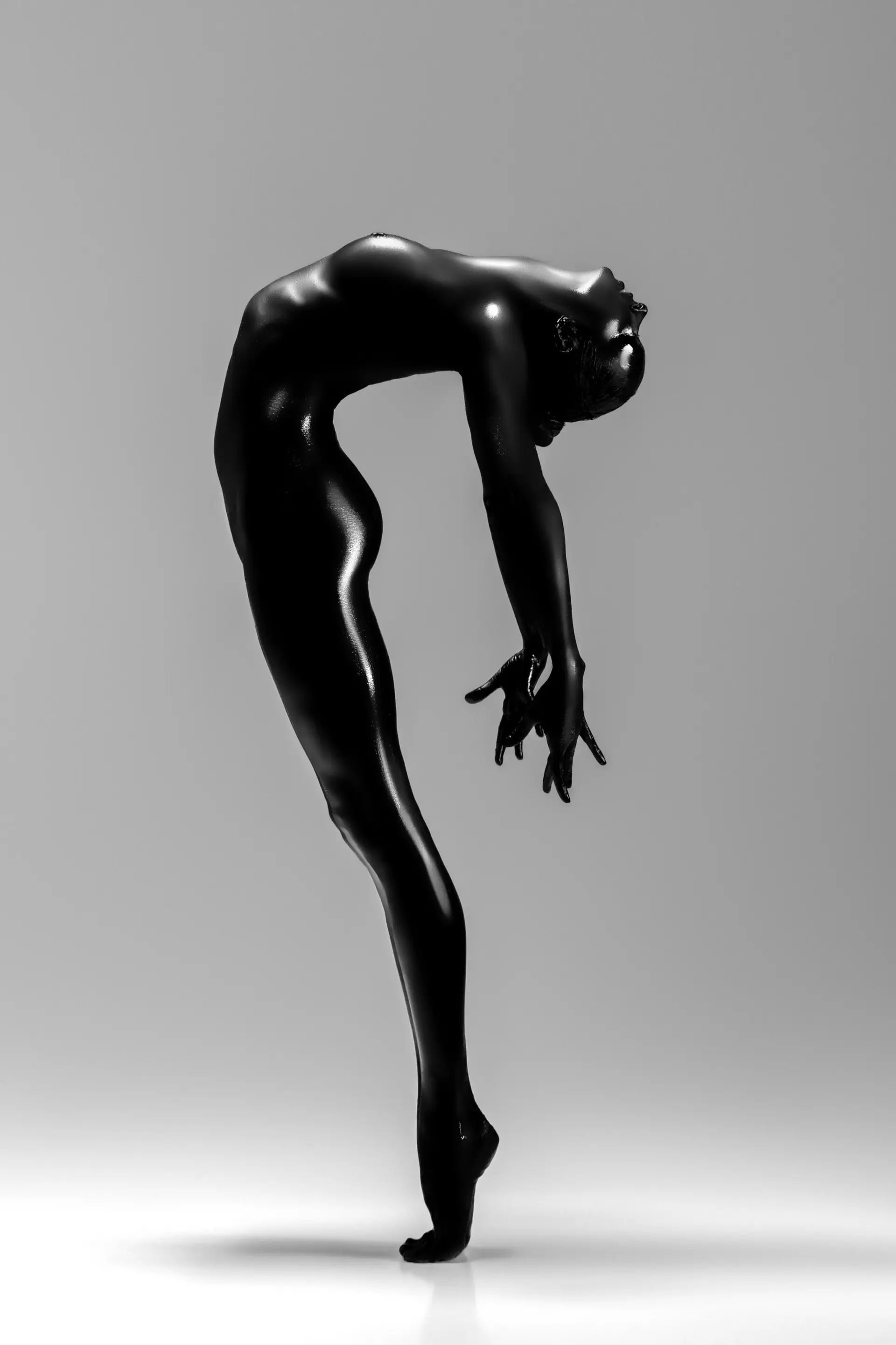 Silhouette of a slender person painted black, standing on toes and bending backward in an elegant, artistic pose against a gray background.