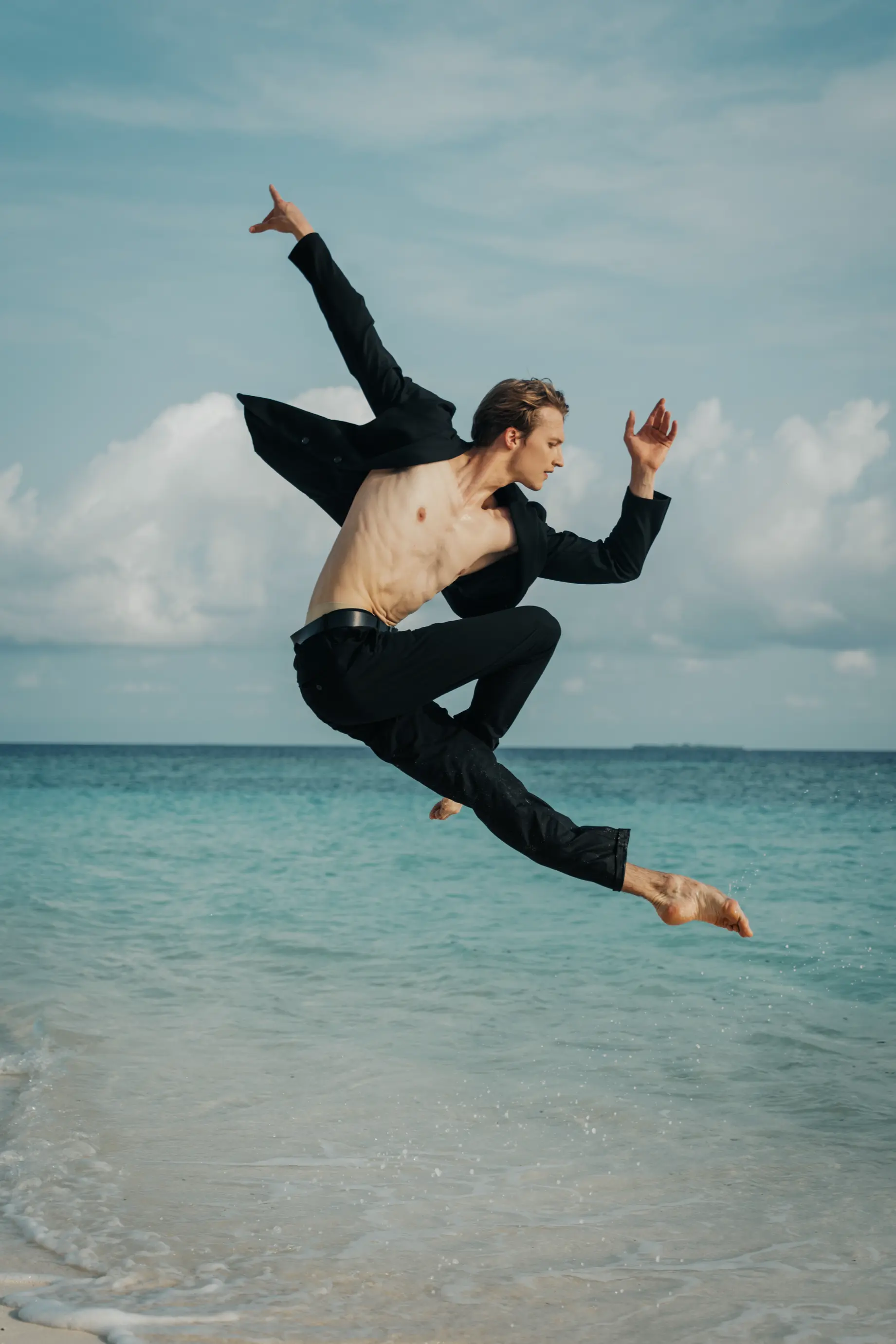 Shirtless man in black pants and jacket mid-jump performing a dynamic dance pose on a beach with blue sea and cloudy sky background.