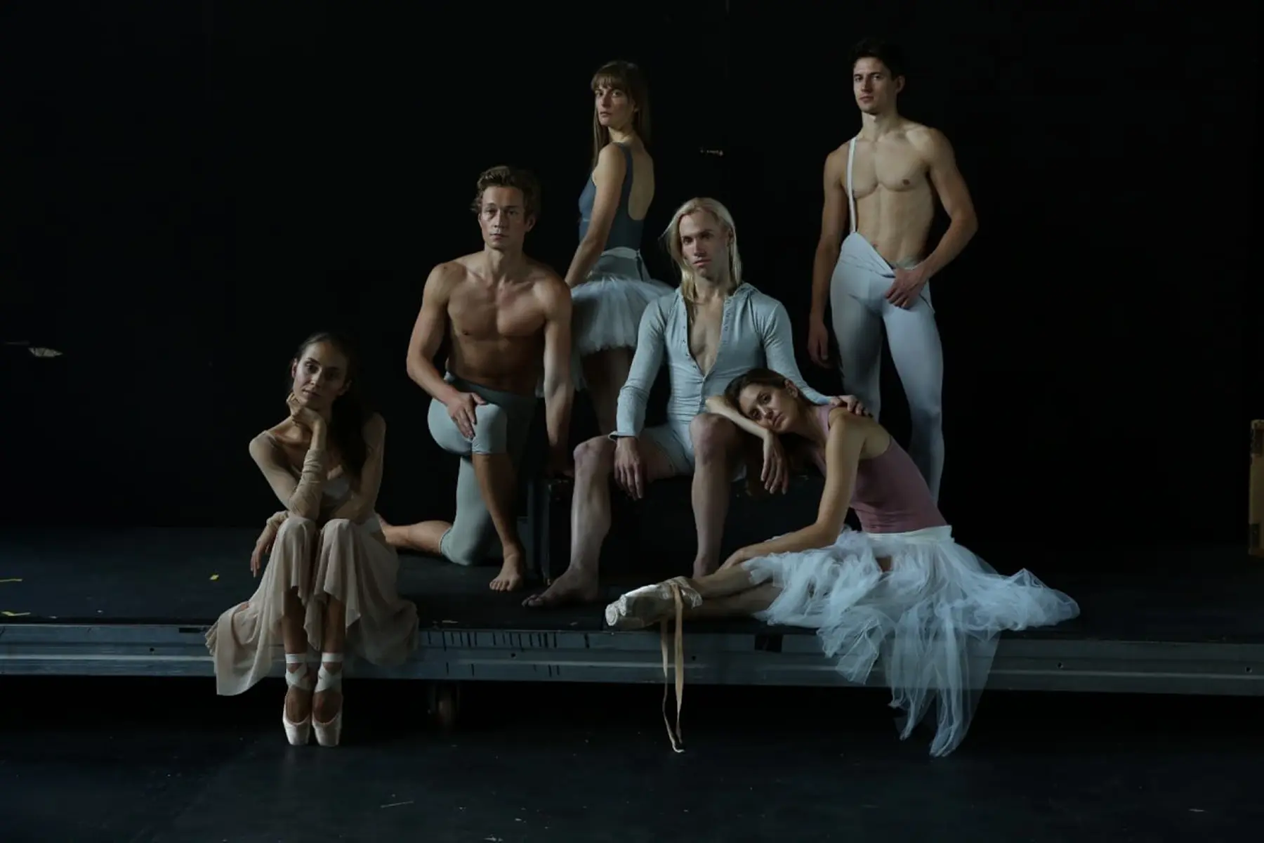 Six ballet dancers posing on stage in varied ballet attire against a black background.
