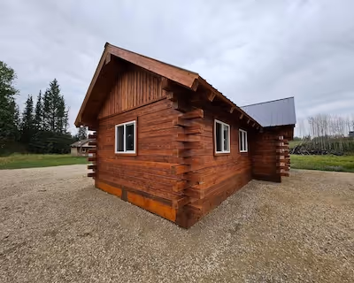 Wooden log cabin exterior with stained siding, small windows, and a gravel yard on an overcast day.