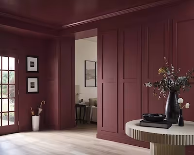 “Elegant interior room with deep burgundy painted walls, decorative paneling, light wood flooring, and a round table with a floral arrangement.