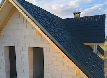 Partially constructed house with white block walls and newly installed dark shingle roof under a clear sky.