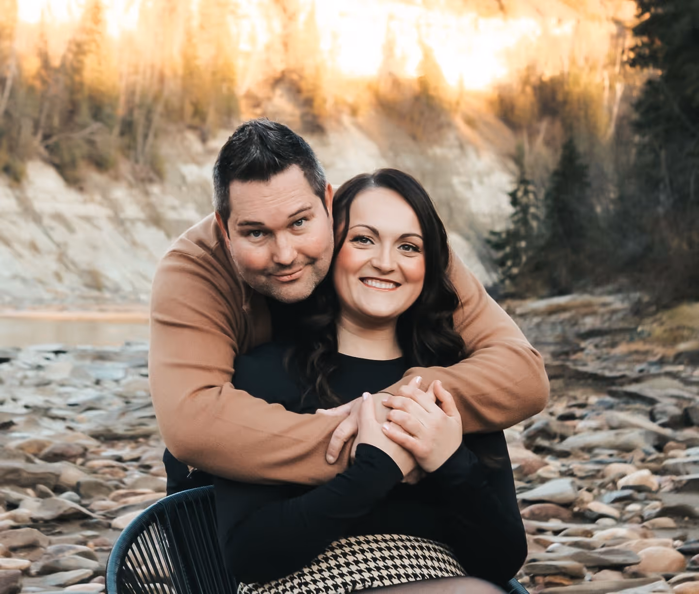 Man in a brown sweater embraces smiling woman in black sitting on a chair outdoors by a rocky riverbank.