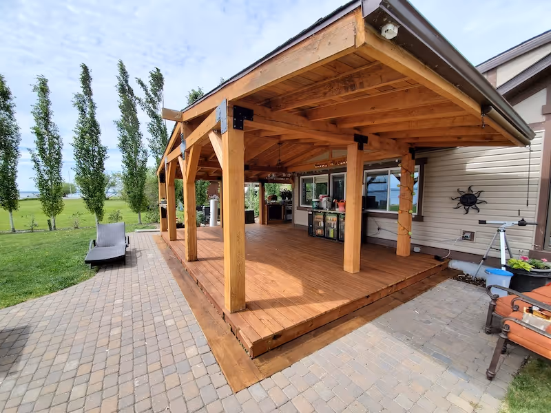 Wooden covered patio attached to a house with brick paver flooring and lawn chairs on the side.
