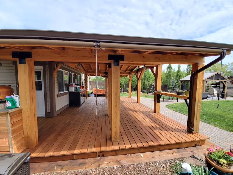 Spacious wooden covered patio with sturdy beams, outdoor furniture, and a green lawn in the background.
