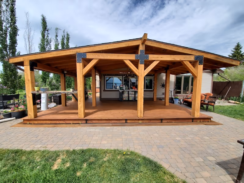Wooden outdoor patio cover structure with a peaked roof over a deck, situated in a backyard with paved flooring and grass.